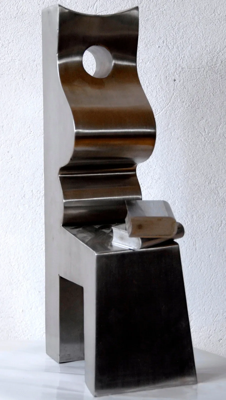 Scultura d’acciaio lucidato: una lastra verticale alta fa da schienale, davanti una superficie ondulata descrive un corpo seduto di profilo con un foro circolare al posto del capo; in basso il piano del grembo con due libri d’acciaio, e sotto un volume trapezoidale che fa da gambe