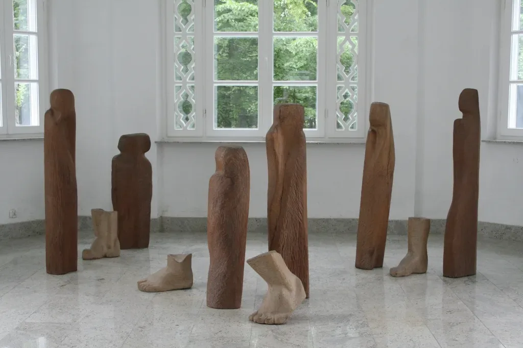 Wygnanie nella sala del museo: sei figure lignee verticali di altezze diverse e quattro grandi piedi di legno posati sul pavimento chiaro, davanti alle grandi finestre