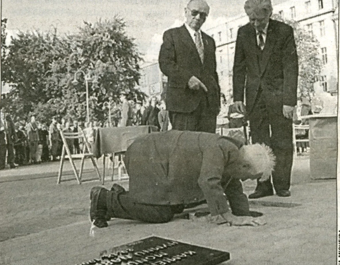 Fotografia di giornale: un uomo lavora inginocchiato sulla lastra della pietra angolare mentre due uomini in abito scuro osservano; in primo piano le lettere di bronzo dell’iscrizione, ancora sciolte