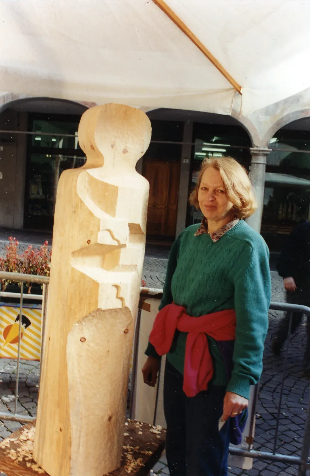 Antonina accanto alla scultura appena finita, sotto il telo bianco della postazione: dietro di lei i portici della piazza, ai piedi del tronco i trucioli. È la 9ª Ex Tempore di Belluno, novembre 1994.