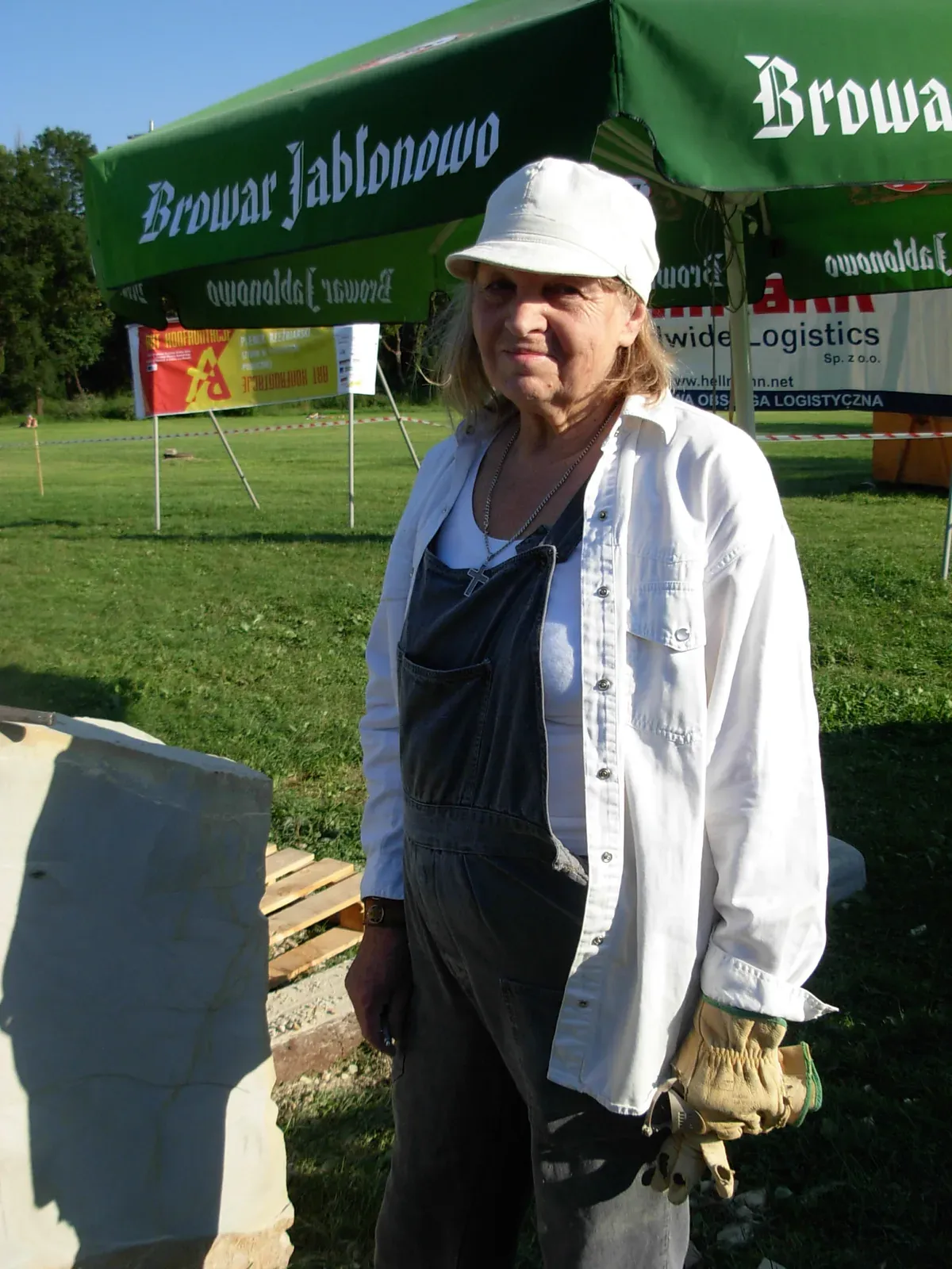 Antonina Wysocka-Jonczak in piedi al plein air, cappello bianco, camicia bianca aperta su una tuta da lavoro scura, i guanti da cava in mano; dietro di lei un gazebo e lo striscione della manifestazione