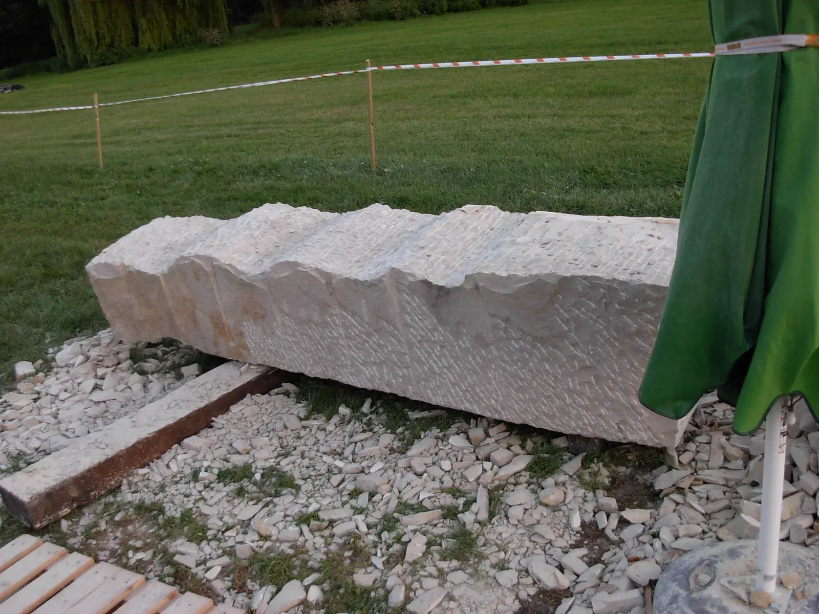 Un lungo blocco di arenaria disteso su travi e pallet nel prato, col bordo superiore già tagliato a onde larghe; attorno, schegge di pietra e il nastro del cantiere