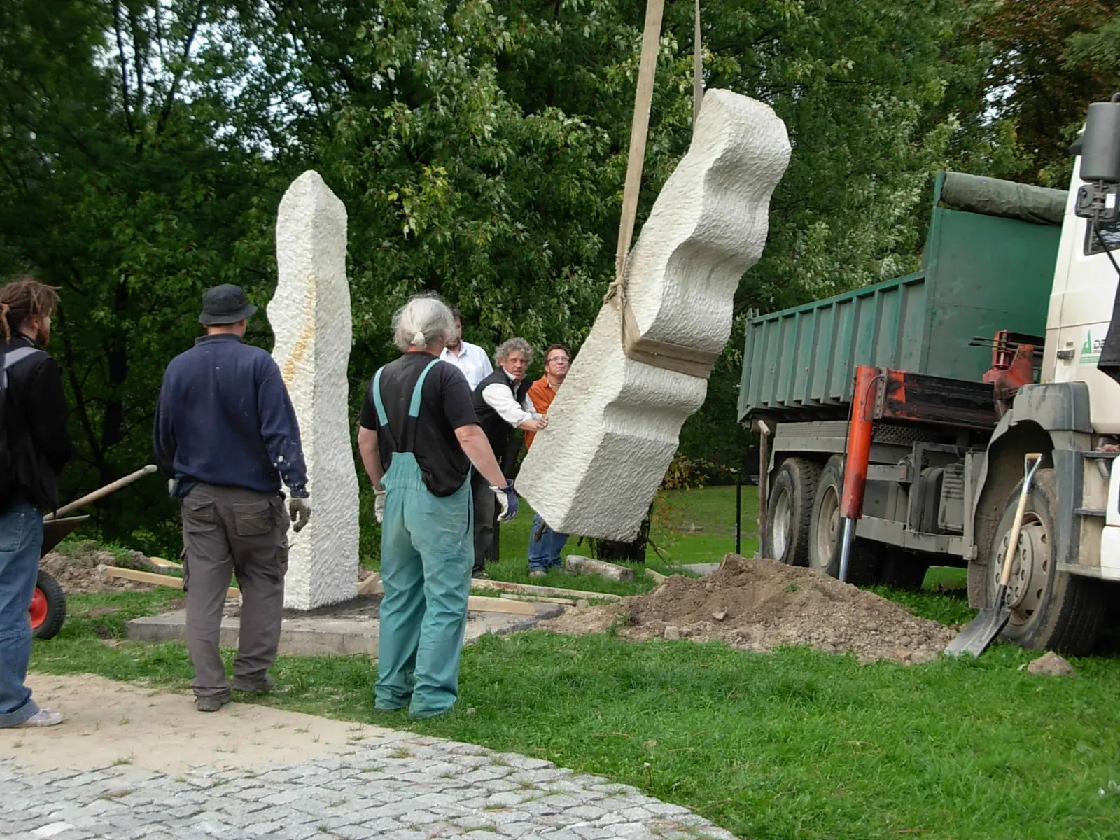 Etiuda — La posa: una stele è già in piedi, l'altra pende dal braccio della gru sopra la buca.