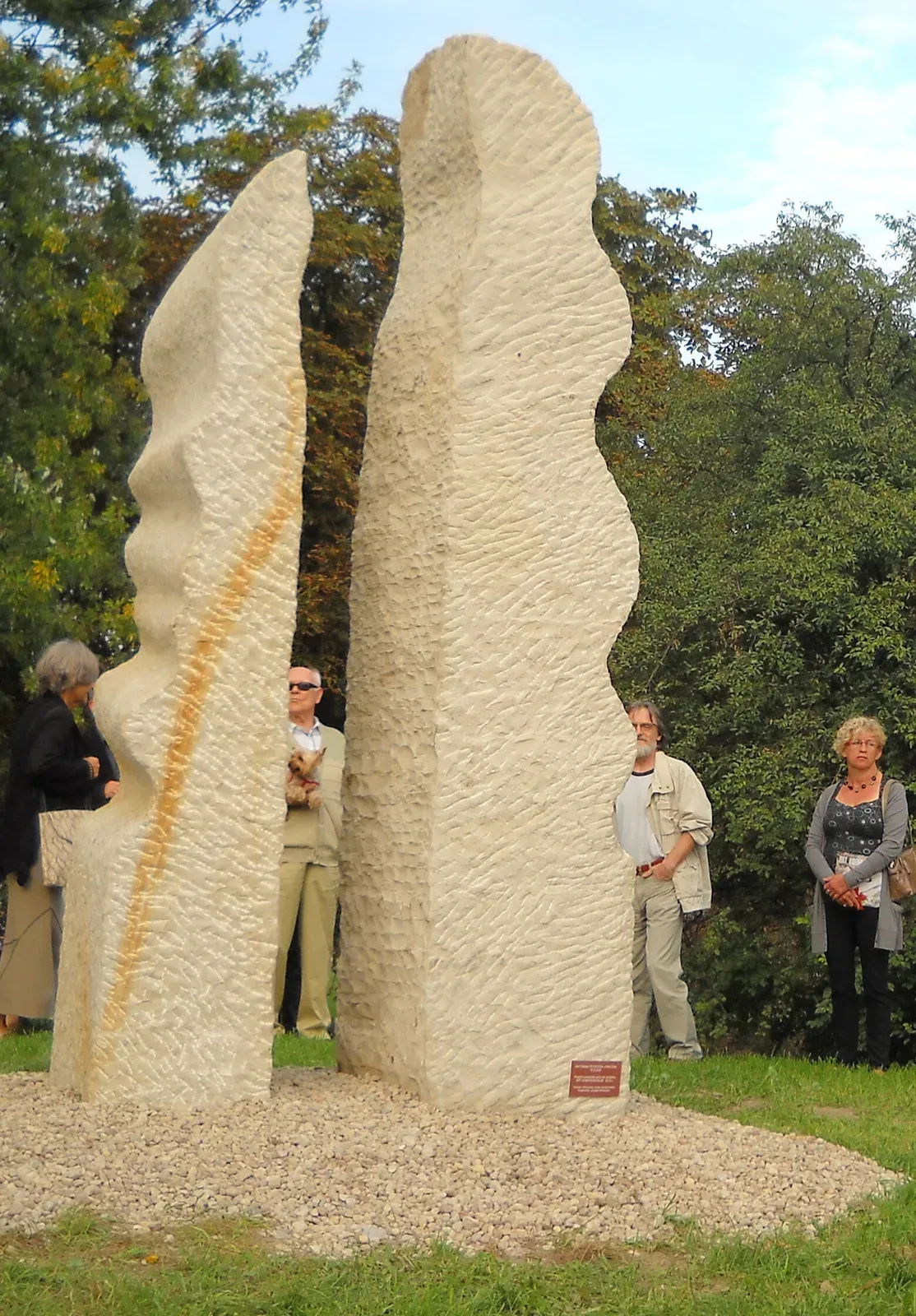 Le due steli in piedi sul letto di ghiaia, viste da vicino: la fessura che le separa, la vena ocra sulla stele di sinistra e la targhetta rossa al piede di quella di destra