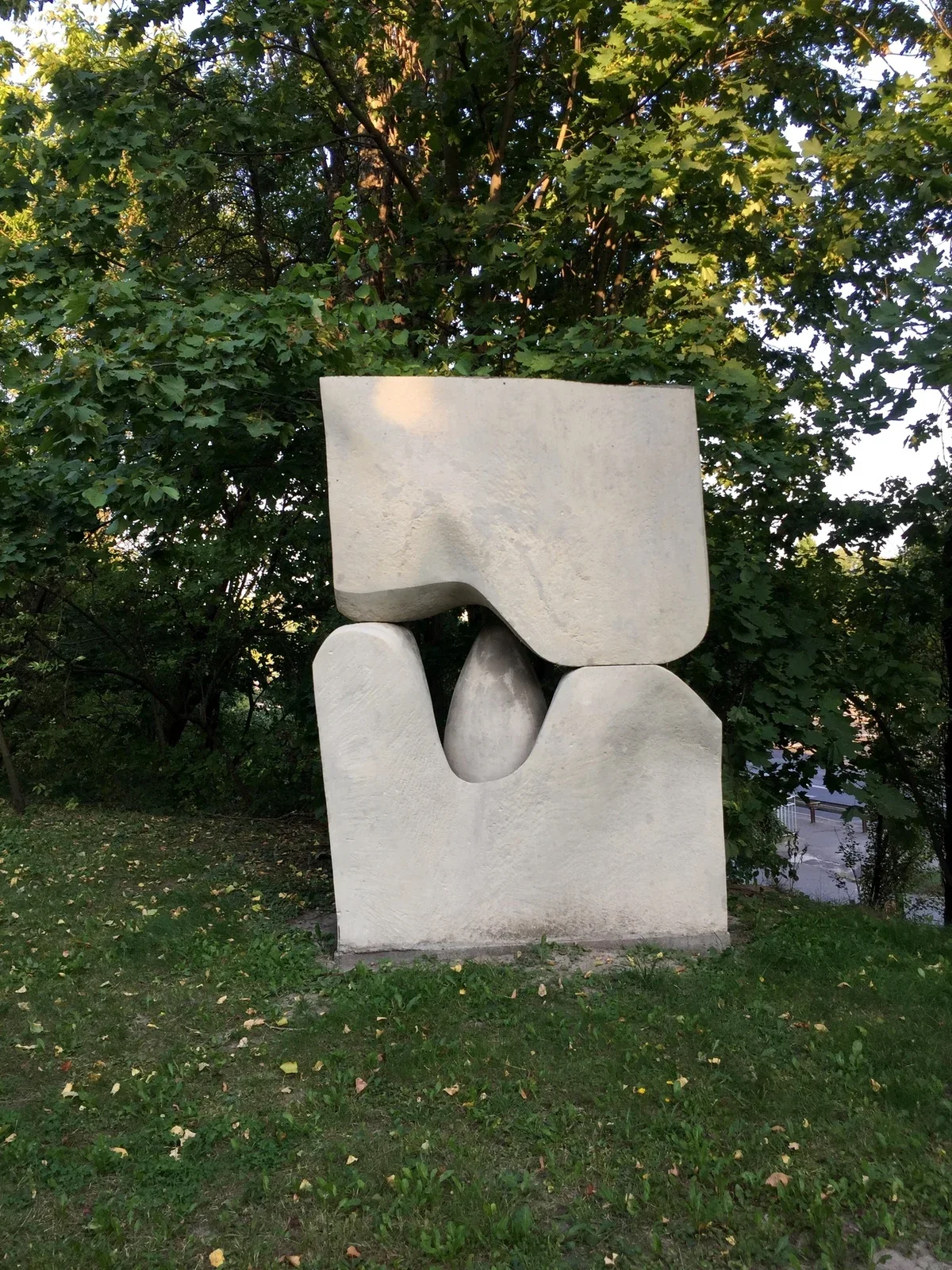 La stessa scultura oggi, a colori: la pietra chiara e pulita in mezzo agli alberi cresciuti, con la strada che si intravede sulla destra