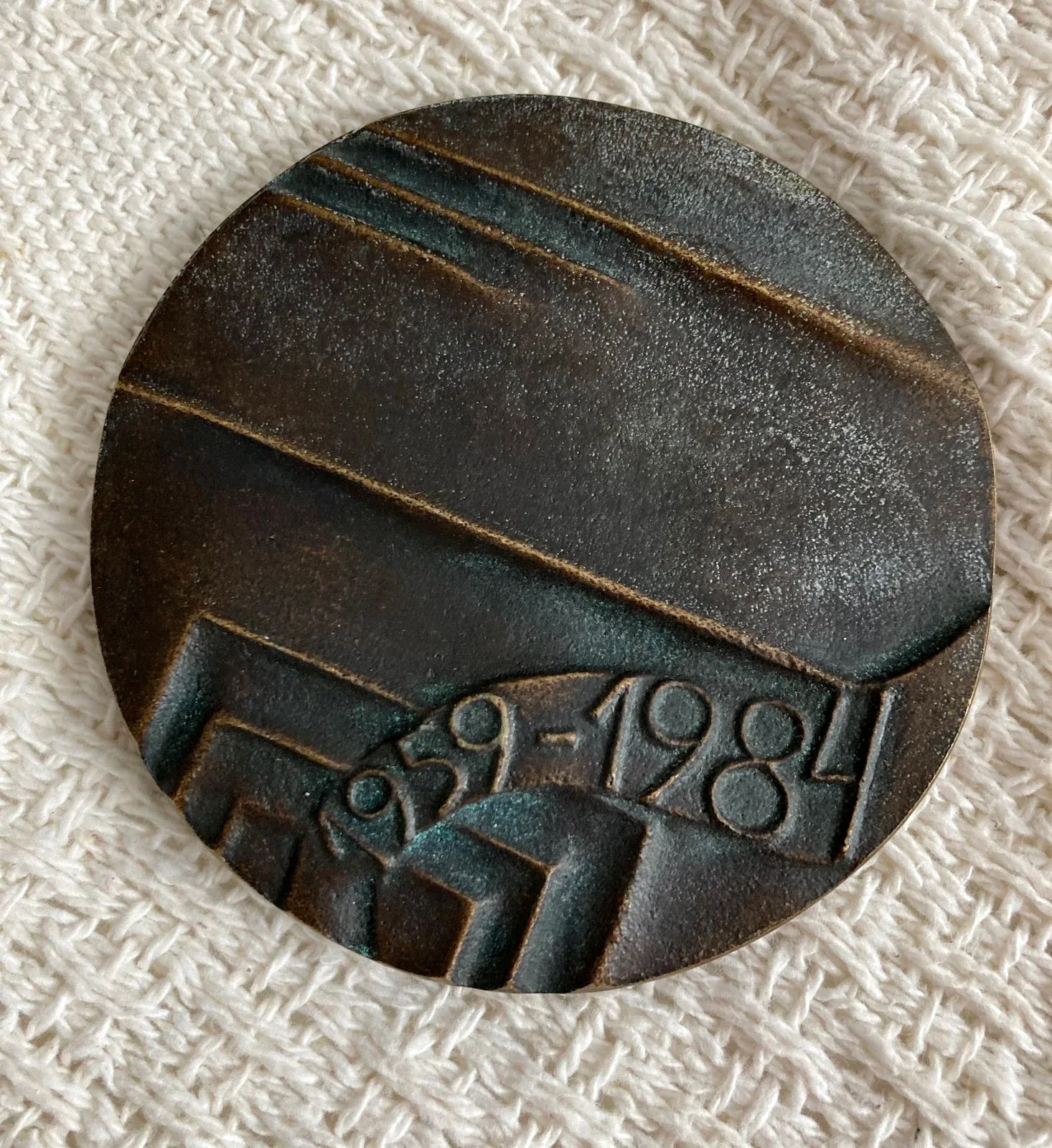 Medaglione di bronzo a patina scura, quasi circolare: la metà superiore è percorsa da lunghe fasce parallele oblique, la metà inferiore da un meandro a gradini quadrati, attraversato da un nastro ondulato su cui corre in rilievo la scritta «1959 - 1984»
