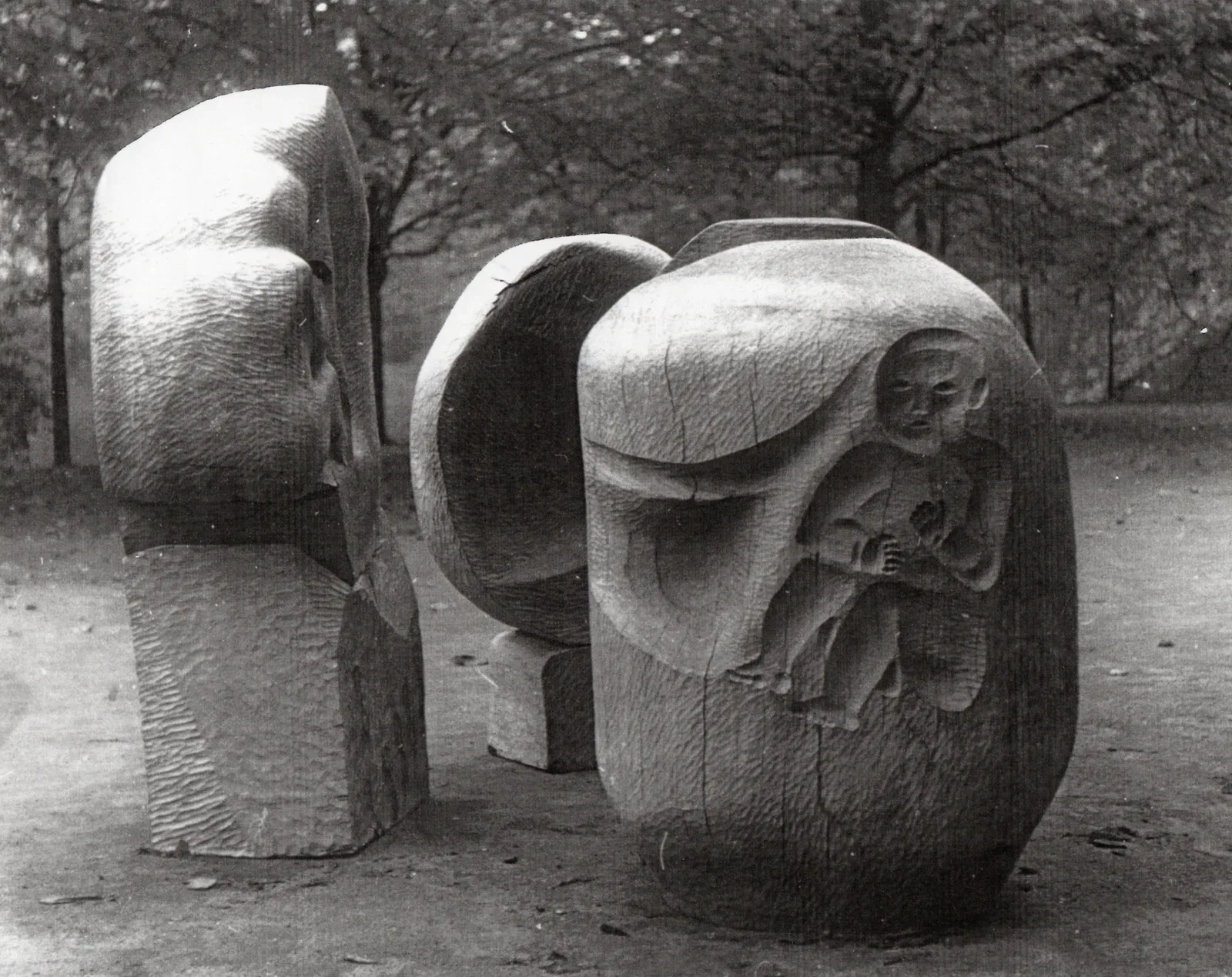 Fotografia in bianco e nero: tre grandi masse di legno arrotondate posate su un vialetto di terra in un parco, con alberi spogli sullo sfondo. Ciascuna è scavata in modo diverso; in quella di destra si apre un vano dentro cui è scolpito un bambino nudo seduto