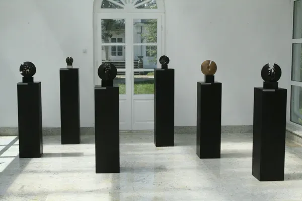 Sei dischi di bronzo su alti plinti neri disposti a semicerchio davanti alla porta a vetri dell’Oranżeria, con il parco alle spalle.