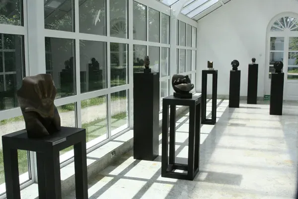 L’Oranżeria in controluce: una fila di plinti neri di altezze diverse lungo le vetrate, con un grande bronzo in primo piano.