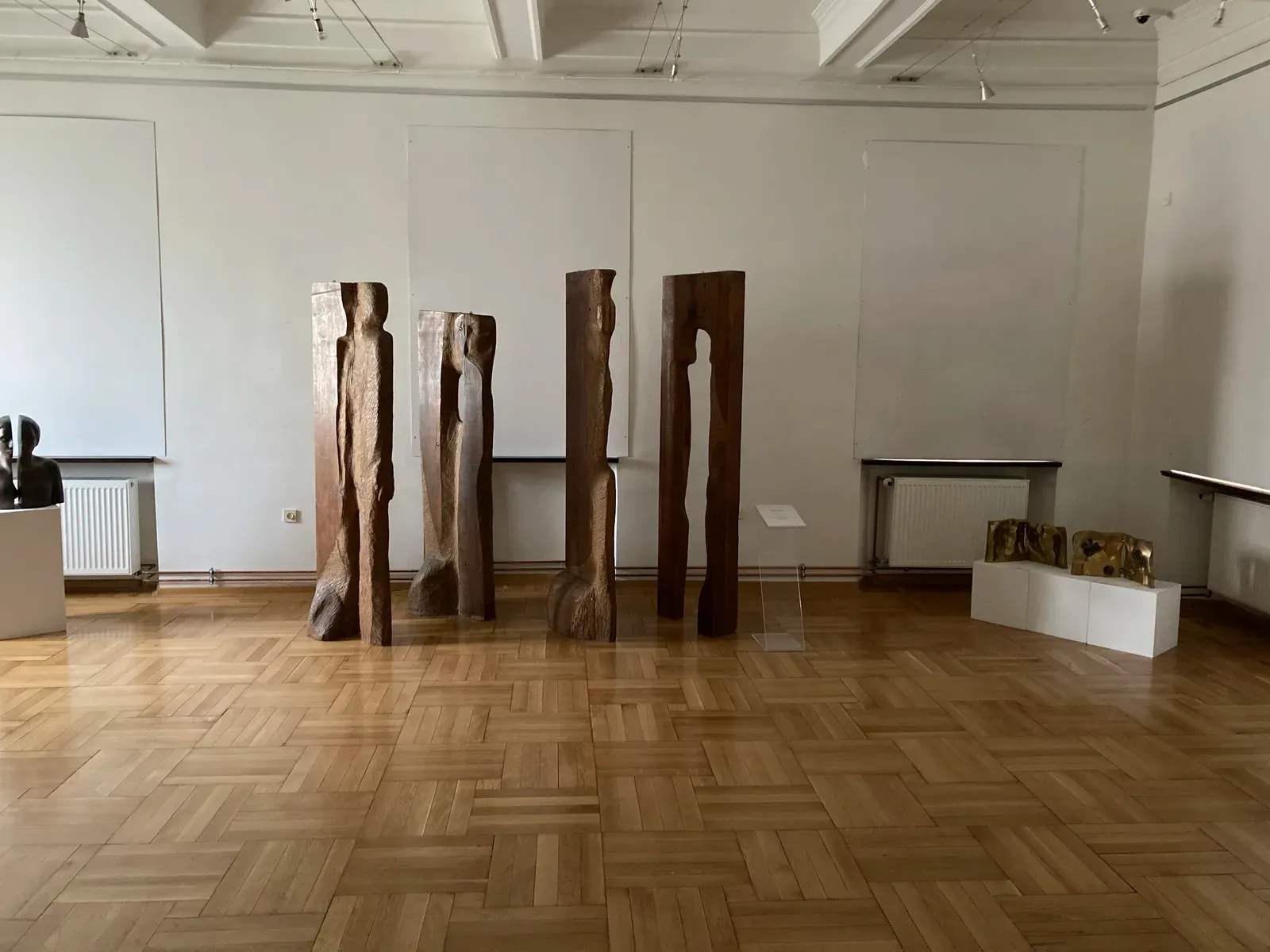 Sala di museo con parquet e quattro alte steli lignee scolpite in fila, piccoli bronzi dorati su plinti a destra