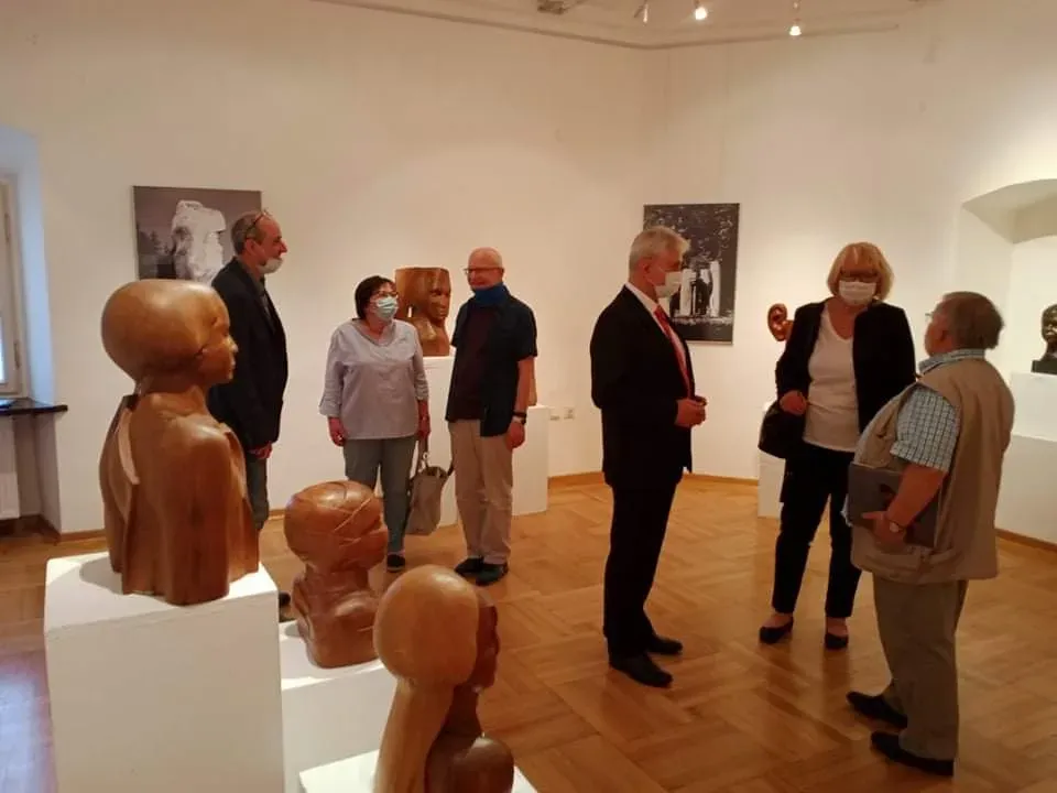 Sala della mostra durante il vernissage: visitatori con la mascherina conversano fra i busti lignei sui plinti bianchi.