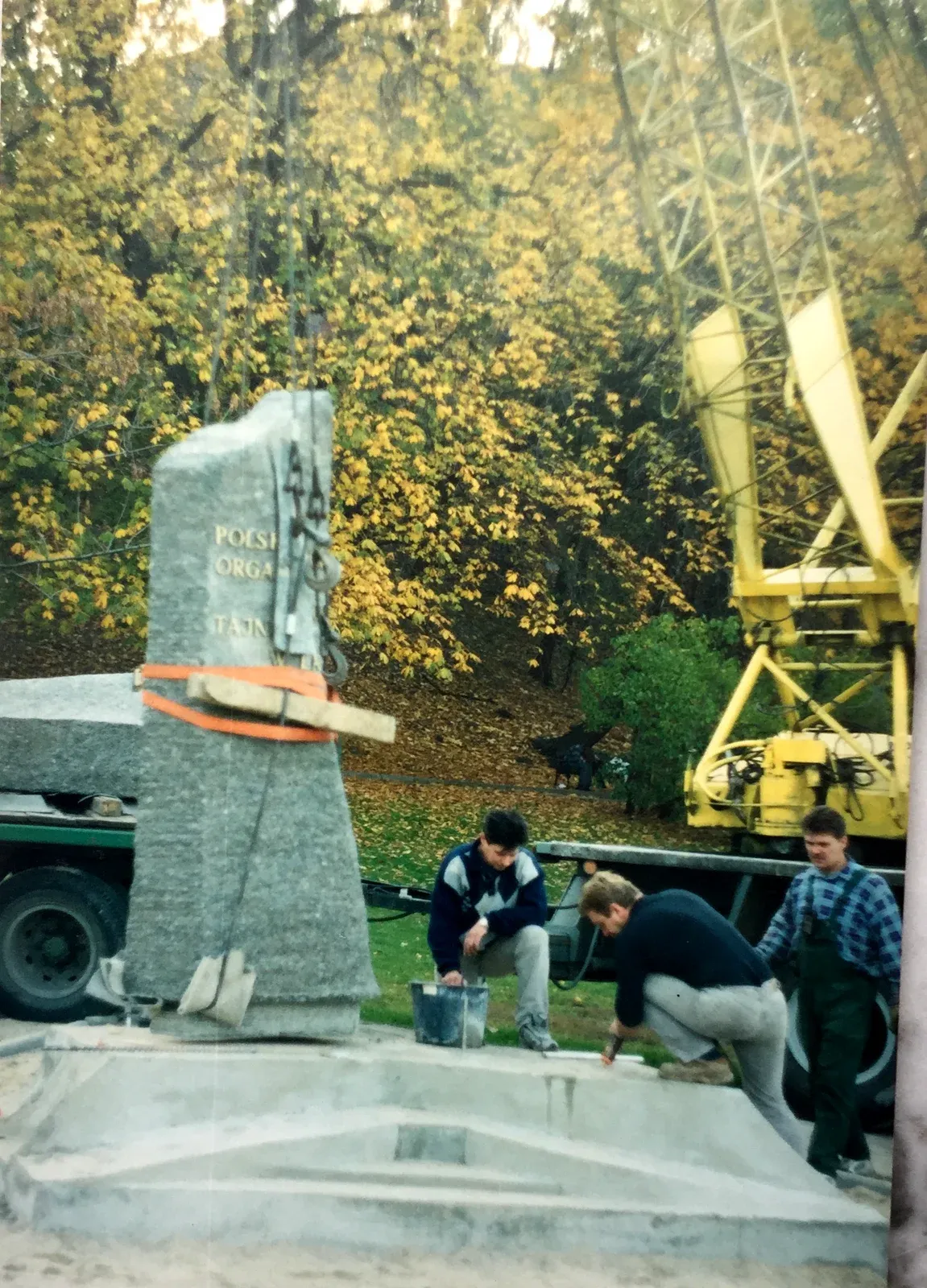 La stele imbragata con cinghie arancioni pende dalla gru sopra la base di cemento, fra gli alberi gialli del parco